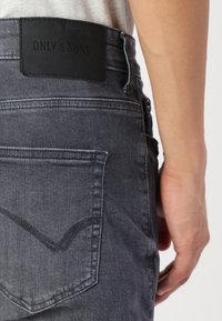 Mørkegrå jeans med et lærremskilt mærket "ONLY & SONS" på taljen, med detaljer på baglommene og et tekstureret stof.