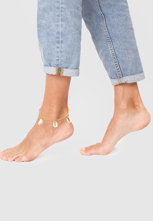 Pulsera de tobillo de oro con acentos de concha marina, llevada en un pie descalzo, combinada con jeans de mezclilla azul claro arremangados.