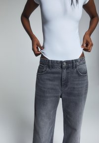 Persona que lleva unos pantalones vaqueros negros desgastados y de gran tamaño, con una camisa blanca ajustada metida por dentro, sosteniendo los bordes de la camisa cerca de la cintura.