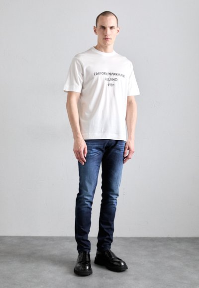 T-shirt en coton blanc avec impression graphique noire, associée à un jean en denim bleu foncé et des chaussures noires à lacets, se tenant sur une surface uniforme.