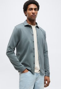 Άνδρας με αφροχτένισμα που φοράει ανοιχτό γκρι πουκάμισο με κουμπιά πάνω από μπεζ μπλουζάκι και ανοιχτό μπλε τζιν, στέκεται με το ένα χέρι στην τσέπη.