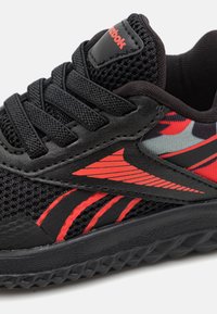 Zapato deportivo negro con parte superior de malla, acentos rojos y suela texturizada. Presenta un parche con el logo y un diseño de franjas laterales audaces.