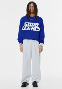 Blauwe gebreide trui met witte tekst "STUD LEGACY", gecombineerd met losse grijze joggingbroek en zwarte schoenen. Lange mouwen en gelaagde manchetten zijn aanwezig.