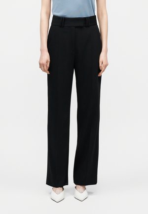 Személy fekete, magasderekú, széles szárú, szabott nadrágot visel, amelyhez öv tartozik, valamint fehér, hegyes orrú cipőt, egyszerű háttér előtt.