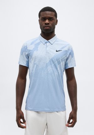 Mann trägt ein hellblaues Nike-Poloshirt mit abstrakten Mustern und weiße Shorts, steht vor einem schlichten weißen Hintergrund.