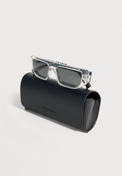Διάφανα ορθογώνια γυαλιά ηλίου με σκούρα φακούς που ακουμπούν σε μια μαύρη σκληρή θήκη με το λογότυπο Dolce & Gabbana σε λευκό φόντο.