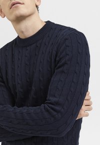 Navy blue knitted sweater with raised cable pattern, ribbed crew neckline, and long sleeves. Fabric appears textured and thick.