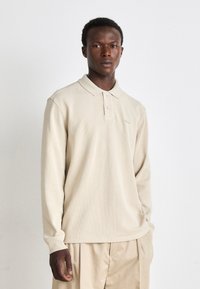 Camisa polo de manga longa bege feita de um tecido texturizado, com um colarinho suave, placket de três botões e um logótipo discreto no peito.