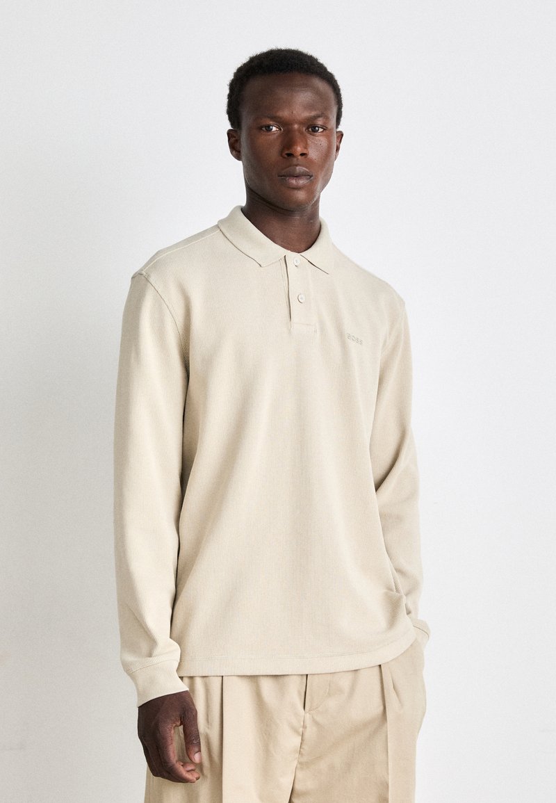 Camisa polo de manga longa bege feita de um tecido texturizado, com um colarinho suave, placket de três botões e um logótipo discreto no peito.