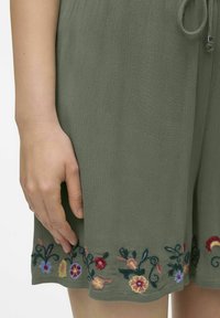 Vestido de tela texturizada color verde oliva con bordado floral en el dobladillo. Presenta una cintura con cordón y un corte holgado.
