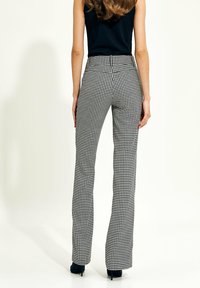 Kobieta w czarno-białych spodniach w wzór houndstooth o prostym kroju i czarnej bluzce bez rękawów, stojąca na jednolitym tle.