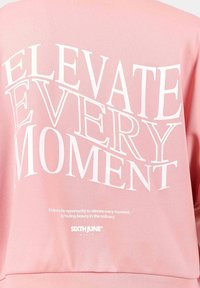 Felpa rosa con grande testo bianco "ELEVATE EVERY MOMENT" e testo più piccolo sotto. Tessuto liscio e design rilassato per un look casual.