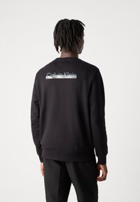 Zwart sweatshirt met een ronde hals, ribgebreide manchetten en een reflecterend "Calvin Klein" logo ontwerp op de achterkant. Gladde stoftextuur.