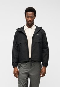 Άνδρας που φοράει μαύρο μπουφάν με φερμουάρ πάνω από μπεζ πλεκτό πόλο και γκρι παντελόνι, που στέκεται μπροστά σε απλό ανοιχτογρίζο φόντο.