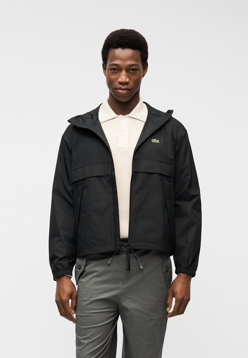 Άνδρας που φοράει μαύρο μπουφάν με φερμουάρ πάνω από μπεζ πλεκτό πόλο και γκρι παντελόνι, που στέκεται μπροστά σε απλό ανοιχτογρίζο φόντο.