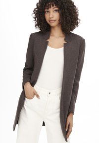 Blazer marrone aperto davanti realizzato in tessuto morbido con design senza colletto. Indossato sopra una maglietta bianca e abbinato a pantaloni bianchi.