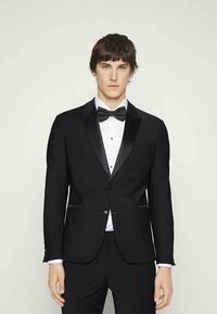 Jovem vestido com um fraque preto com lapelas de cetim, camisa de vestir branca e gravata-bow preta, em pé diante de um fundo claro e liso.