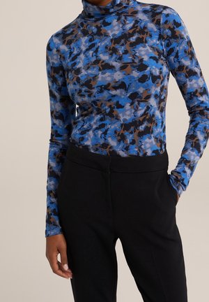 Femme portant un haut à manches longues et col montant avec un imprimé abstrait bleu et marron, et un pantalon noir ajusté, une main dans la poche.