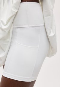 Κοντινό πλάνο ενός ατόμου που φοράει λευκό ποδηλατικό σορτς με ψηλή μέση και πλαϊνή τσέπη, καθώς και λευκό τοπ με τύπου ντραπέ κομμάτι.