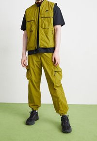 Man i olivgröna cargobyxor och matchande väst över en svart t-shirt, med svarta sneakers, står på grön golv.