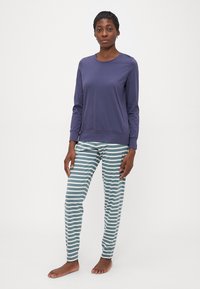 Kvinna som står barfota och bär en långärmad lila topp och teal-och-vita randiga pyjamasbyxor framför en enfärgad bakgrund.