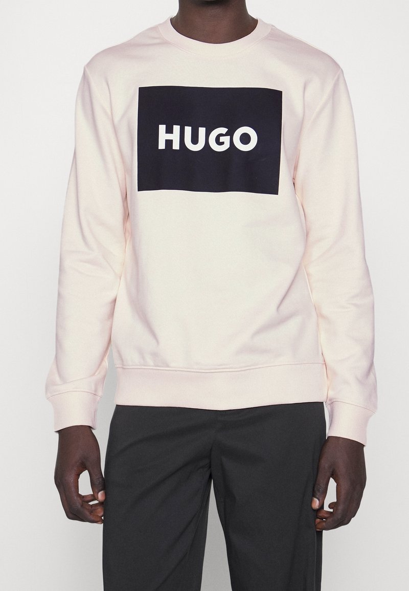 Personne portant un sweat-shirt à manches longues rose clair avec un logo rectangulaire noir et le texte « HUGO », assorti à un pantalon noir.
