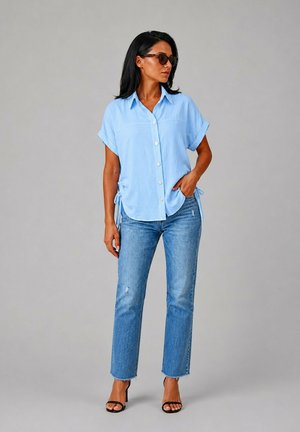 Femme portant des lunettes de soleil, une chemise bleu clair à manches courtes boutonnée, un jean bleu et des talons ouverts noirs, debout devant un fond gris.