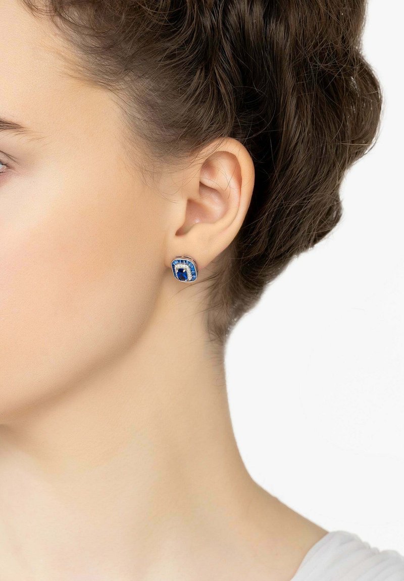 Orecchini a stud con gemme blu di forma rettangolare, dettagli in argento e un bordo testurizzato, mostrati su un orecchio.