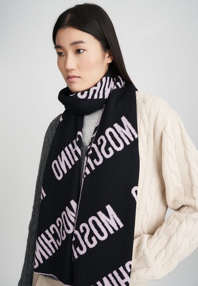 Fiatal nő egyenes fekete hajjal, színes blokkokból készült kötött pulóvert és fekete sálat visel, amelyen ismétlődő rózsaszín "MOSCHINO" felirat található.
