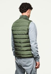 Vihreä puhviliivi, jossa on tikatut paneelit, korkea kaulus ja sileä pintakuvio. Päällä vaalea harmaa pitkähihaisen collegepaita.