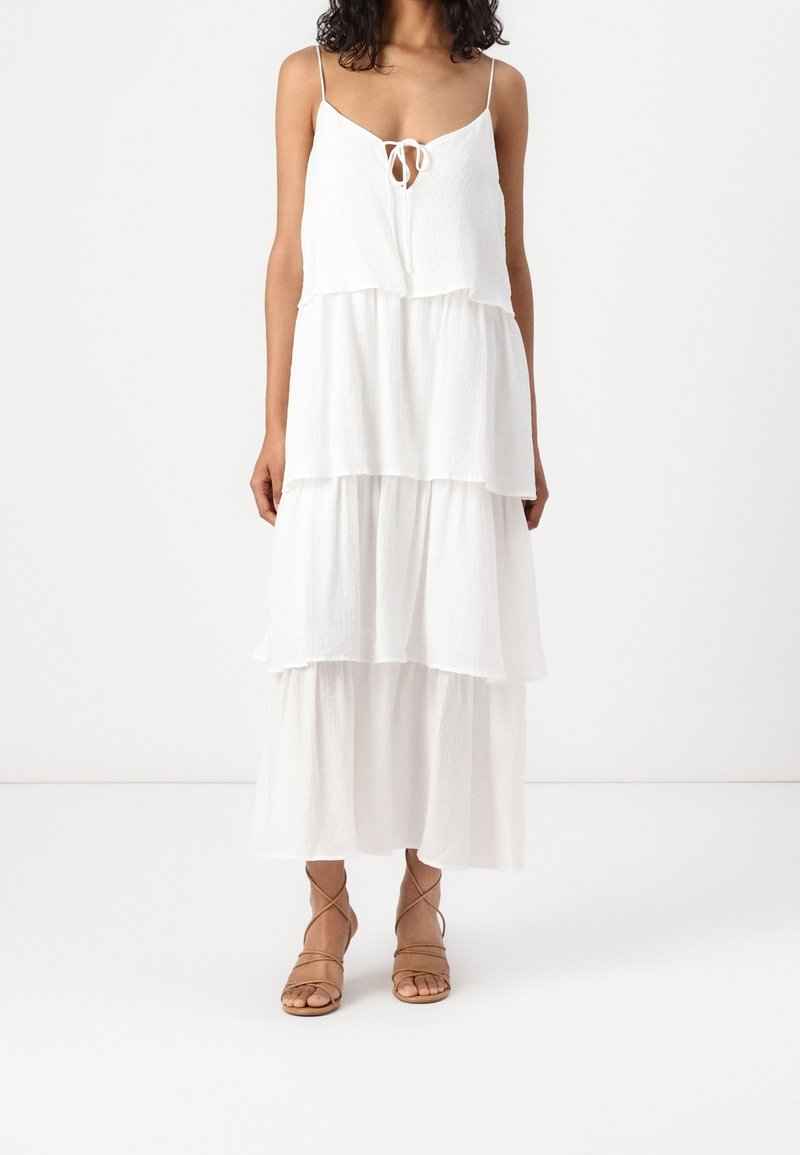 Femme portant une robe maxi blanche à volants avec fines bretelles et détails lacés, associée à des sandales à talons hauts marron clair à brides.