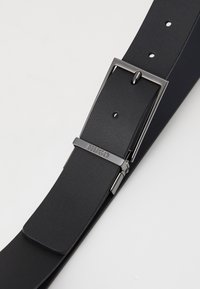 Ceinture en cuir noir avec une texture lisse, une boucle en métal argenté et un accent logo. Le design comprend plusieurs trous d'ajustement.