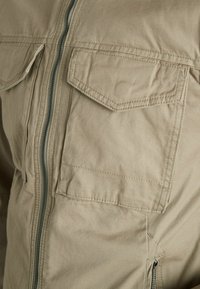 Beige jacket featuring a front zipper, two chest pockets with flaps, and textured fabric. Straight cut with reinforced stitching details.