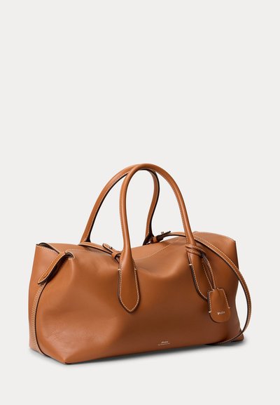 Hellbraune Lederreisetasche mit doppelten Griffen, sichtbaren Nähten und einem kleinen angebrachtem Etikett, auf hellem, einfarbigem Hintergrund platziert.