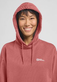 Roze hoodie van zacht materiaal met een trekkoord in de capuchon. Bevat een klein wit logo op de borst. Gladde textuur, casual ontwerp.