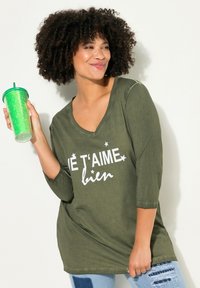Camiseta de manga 3/4 en color verde oliva con escote en forma de v, que presenta el texto en blanco "J'É T'AIME bien" y acentos de estrellas. Combinada con una taza de purpurina verde.