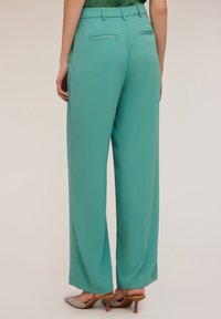 Vida byxor i teal tyg, med hög midja, bakfickor och en mjuk yta, parat med lätta, metalliska klackskor.
