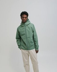 Fiatal férfi, zsályazöld kapucnis dzsekiben és bézs nadrágban, egy egyszínű szürke háttér előtt áll, egyik kezét a zsebében tartva.