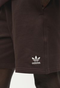 Σκούρο καφέ σορτς Adidas με λευκό κεντημένο λογότυπο τριφύλλι κοντά στο τελείωμα, φορεμένα από άτομο με σκούρο δέρμα.