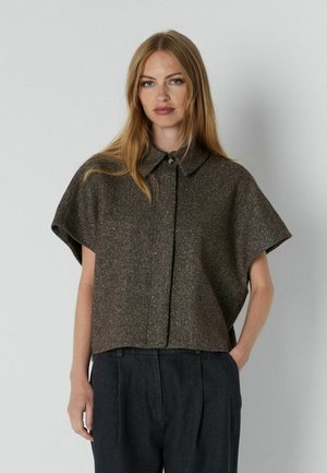 Un mannequin se tient devant un fond gris uni, vêtu d’une veste marron à manches courtes et à col, ainsi que d’un pantalon foncé.