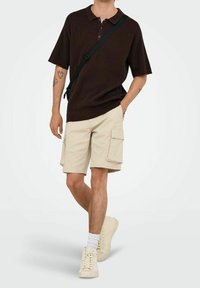 Brązowy dzianinowy polo, beżowe krótkie spodenki cargo z bocznymi kieszeniami oraz kremowe sneakersy w połączeniu z białymi skarpetkami, stojące na neutralnym tle.