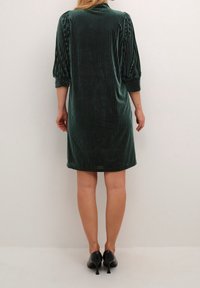 Vestido de terciopelo verde oscuro con rayas verticales, mangas abullonadas de tres cuartos y dobladillo redondeado, combinado con zapatos de tacón alto negros.