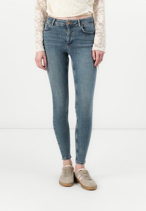 Jean en denim bleu skinny avec une taille mi-haute, un délavage subtil et un ourlet effiloché, associé à des baskets en daim beige.