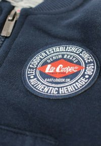 Veste zippée en polaire bleu marine avec un logo rond brodé comportant des accents rouges et blancs, indiquant "Lee Cooper Established Since 1908".