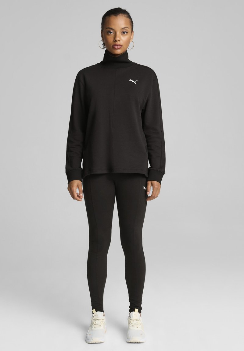 Sudadera negra con cuello alto y logo bordado, combinada con leggings negros y zapatillas deportivas blancas. Diseño sencillo y ajustado.