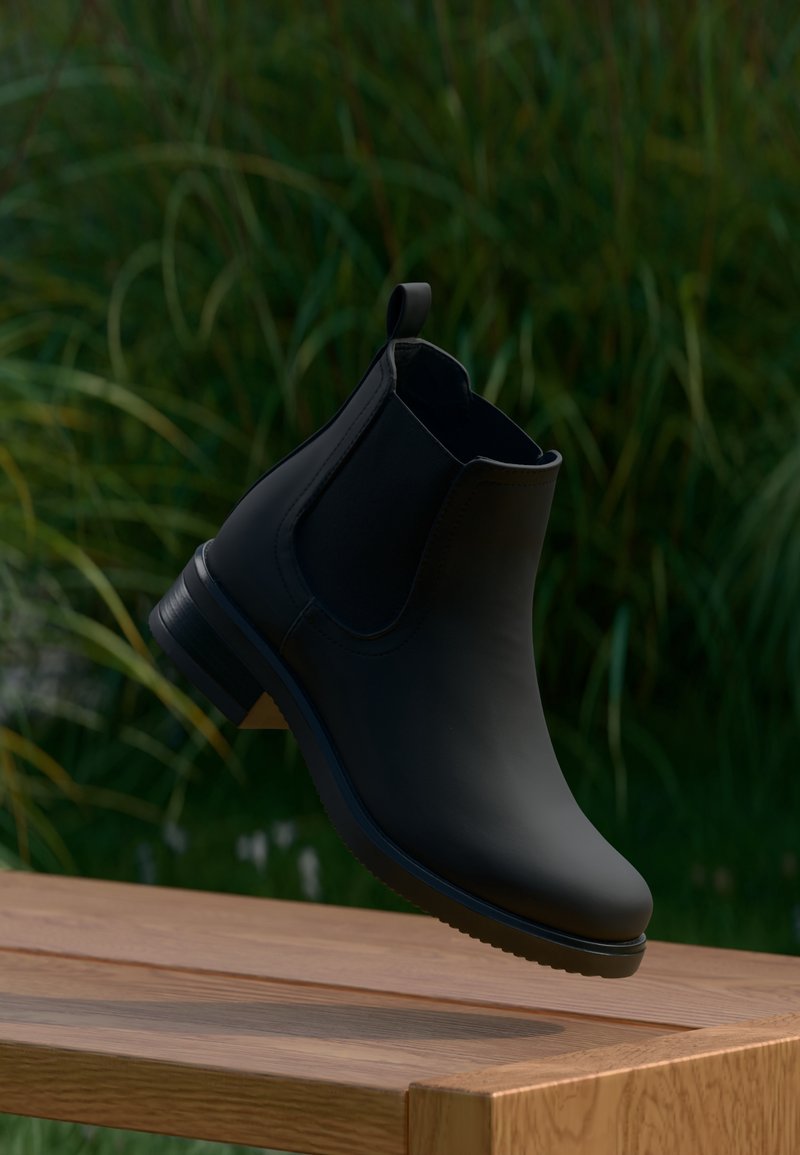 Botte Chelsea en cuir noir flottant au-dessus d’un banc en bois avec des plantes vertes floues en arrière-plan.