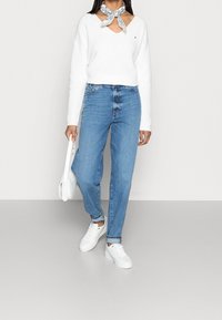 Witte geribde trui met V-hals, blauwe spijkerbroek met een gemiddelde wassing, witte sneakers en een blauwe gebreide sjaal om de nek geknoopt.