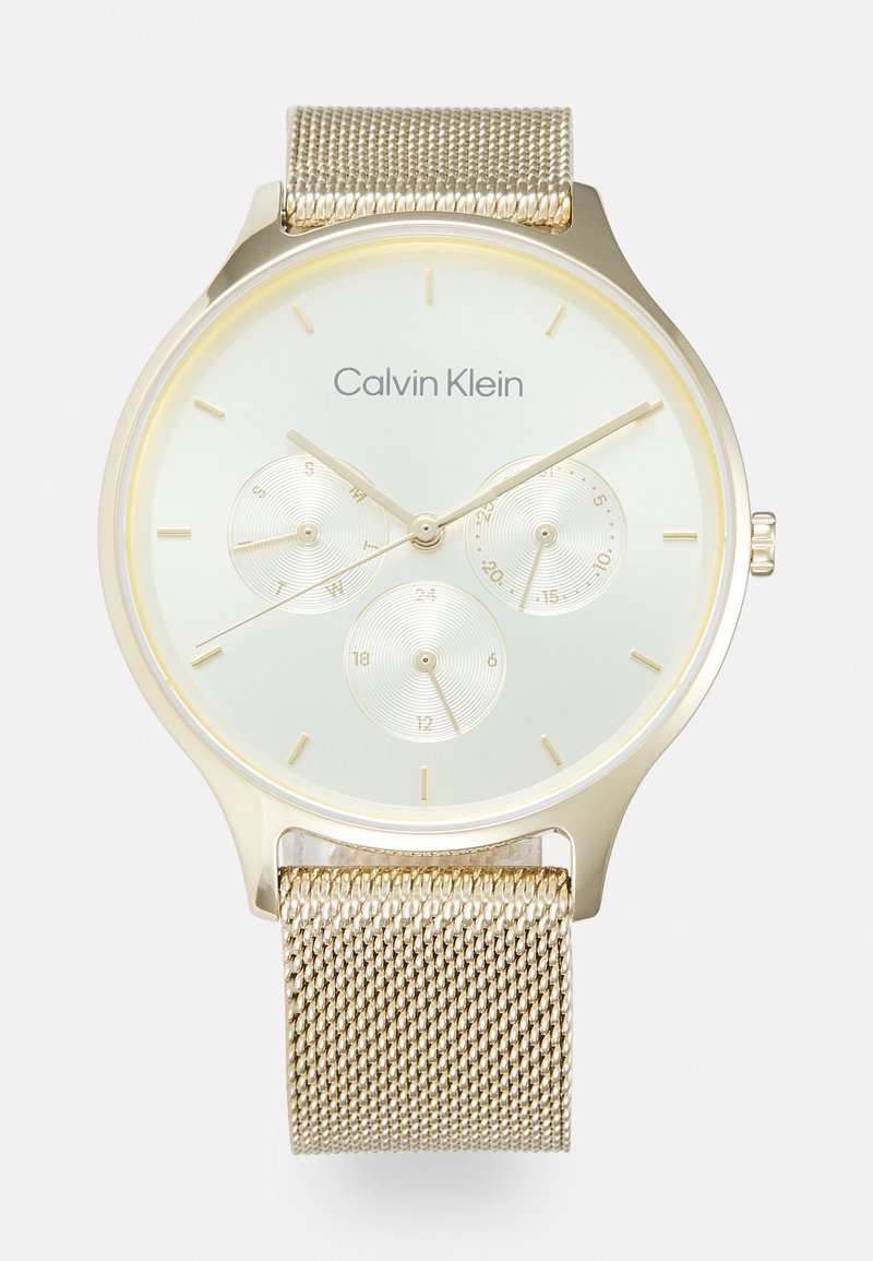 Arany színű óra hálós pánttal, világoszöld számlappal, három alsó számlappal, minimalista óramutatókkal és a Calvin Klein logójával a tetején.
