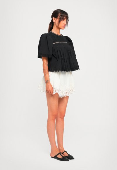 Woman with brown hair in a ponytail wearing a loose black top, white lace shorts, and black pointed slip-on shoes, standing against a plain background.