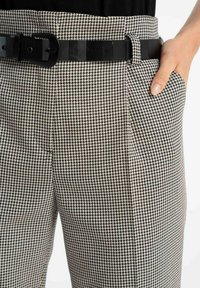 Zbliżenie na czarno-białe spodnie w wzór houndstooth z czarnym paskiem, na którym widać rękę spoczywającą w kieszeni spodni.